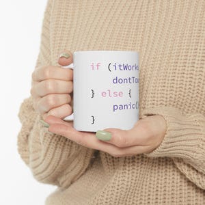 May include: White ceramic mug with the text "if (itWorks dontTouch) } else { panic() }" in a purple and black font. The mug is held by a person wearing a beige sweater.