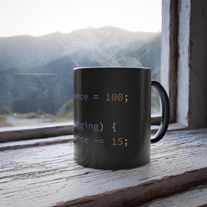 Peut inclure: Mug noir avec de la vapeur blanche. Le mug présente du code de programmation en texte blanc et violet. Il est posé sur une surface en bois blanc, avec une vue panoramique sur les montagnes en arrière-plan.