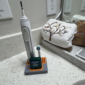Pode incluir: Uma escova de dentes elétrica branca e prateada numa base de plástico cinzenta e laranja com padrão de favo de mel. A base contém um pequeno recipiente de fio dentário verde Oral-B Scope e uma cabeça de substituição ao lado do cabo.