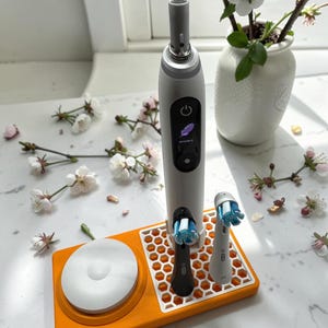 Puede incluir: Un cepillo de dientes eléctrico blanco y gris con dos cabezales y una base de carga, colocado en una bandeja naranja con un patrón de panal. El cepillo de dientes está junto a un pequeño jarrón con flores blancas.