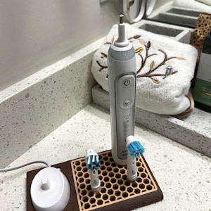 Könnte beinhalten: Eine weiße und graue elektrische Zahnbürste mit zwei Bürstenköpfen und einer Ladestation. Die Zahnbürste und die Bürstenköpfe befinden sich auf einem braunen und beigefarbenen Tablett mit Wabenmuster. Die Ladestation ist weiß und rund.