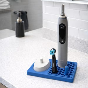 May include: A grey electric toothbrush with a black power button and a blue and white brush head. It sits on a blue silicone tray with a charging base and a spare brush head. Bathroom countertop setting.