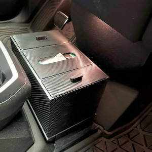May include: A black rectangular car console organizer with a carbon fiber texture. The unit features two small storage compartments with pull tabs and a central slot for dispensing tissues, designed to fit securely within a vehicle interior.