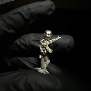 Puede incluir: Pequeña figura plateada de una persona sosteniendo un rifle AK-47. La figura está detallada con un casco y equipo táctico. La figura está montada sobre una pequeña base, sobre un fondo oscuro.