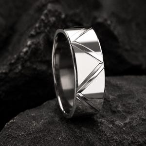Könnte beinhalten: Ein silberner Ring mit geometrischem Design. Der breite Ring hat eine Reihe diagonaler Linien, die ein Muster bilden. Der Ring wird auf einer dunklen, strukturierten Oberfläche präsentiert, was seine polierte Oberfläche hervorhebt.