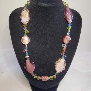 May include: A colorful necklace with a variety of beads and glass elements. The necklace features large, irregularly shaped purple and cream-colored glass pieces, interspersed with smaller, round beads in shades of pink, green, blue, and gold. The necklace is displayed on a black jewelry stand.