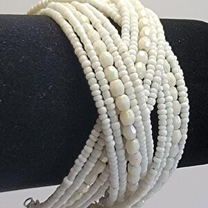 May include: A multi-strand bracelet composed of numerous rows of small, white, opaque seed beads interspersed with larger, faceted, iridescent white beads. The strands are arranged in a crisscross pattern, creating a layered, textured appearance.