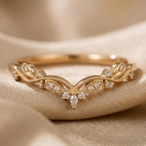 May include: A gold-colored ring with a curved design, featuring small, sparkling gemstones. The ring has a delicate, ornate pattern with leaf-like details and is set against a soft, neutral background.