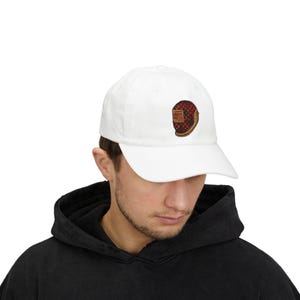 Può includere: Cappellino da baseball bianco con un colorato design ricamato di un casco da saldatura. Il design del casco presenta colori rosso, marrone e oro. Il cappellino è realizzato in un materiale morbido e ha una visiera curva.