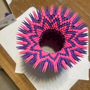 May include: A handmade, geometric paper sculpture in shades of pink and purple. The sculpture has a circular shape with a central opening and a spiky, textured surface. It sits on a white sheet of paper on a wooden table.