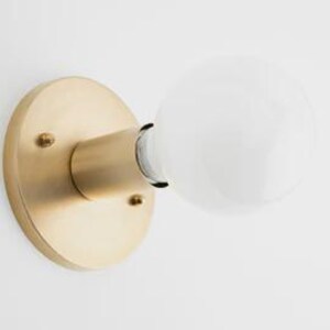 May include: A brass-colored wall sconce with a round base and a cylindrical arm holding a white globe light bulb. The sconce is mounted on a white wall, showcasing a minimalist design.