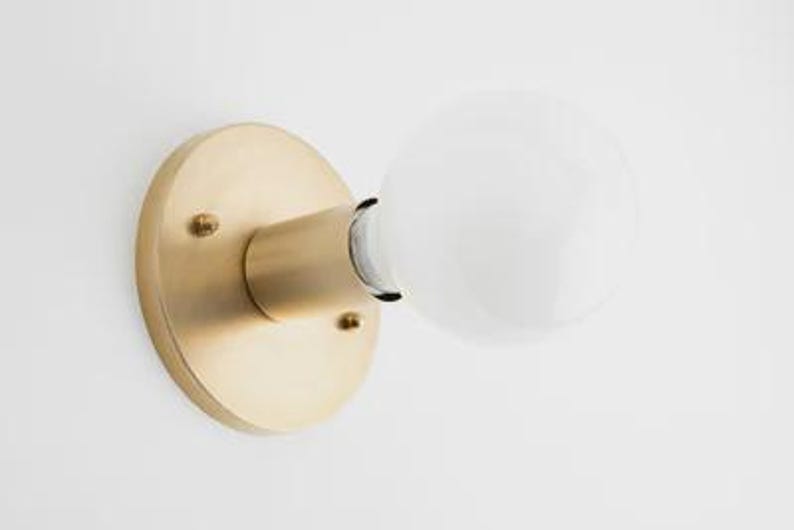 May include: A brass-colored wall sconce with a round base and a cylindrical arm holding a white globe light bulb. The sconce is mounted on a white wall, showcasing a minimalist design.