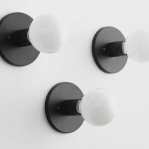 May include: Three black wall sconces with white globe-shaped light bulbs. Each sconce has a circular base and a short arm extending to hold the bulb. The sconces are mounted on a white wall, creating a minimalist design.