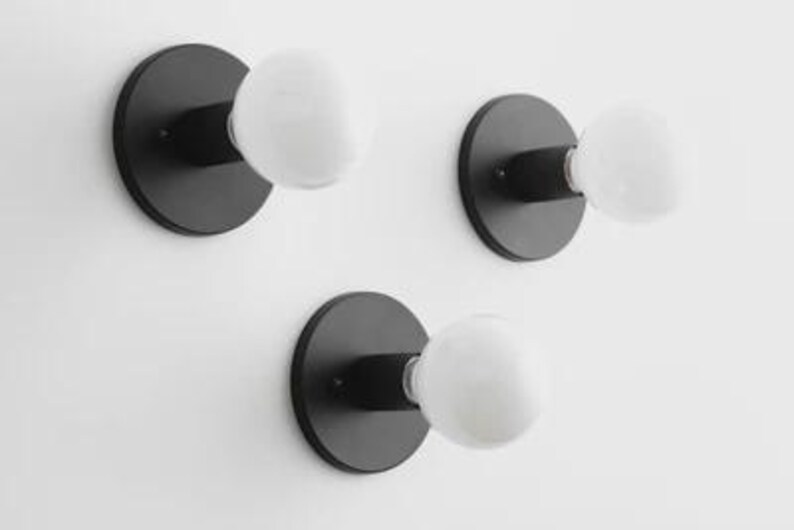 May include: Three black wall sconces with white globe-shaped light bulbs. Each sconce has a circular base and a short arm extending to hold the bulb. The sconces are mounted on a white wall, creating a minimalist design.