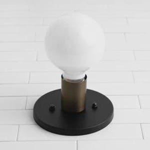 May include: A modern wall sconce featuring a large, round, white light bulb. The bulb is set in a brass-colored cylinder, which is mounted on a black, circular base. The fixture is designed for indoor use.