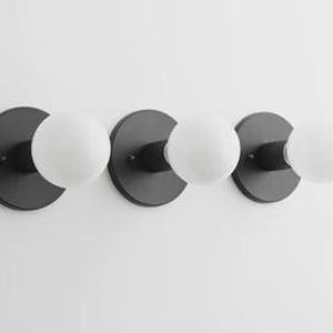 May include: A modern, three-light fixture with a minimalist design. The fixture features three matte black circular backplates, each supporting a spherical, frosted white glass bulb. The bulbs are evenly spaced, creating a balanced and contemporary aesthetic.