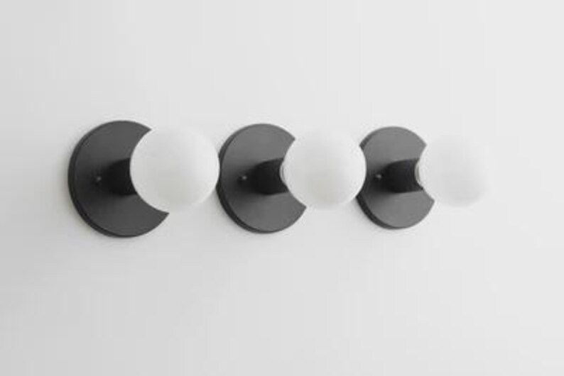 May include: A modern, three-light fixture with a minimalist design. The fixture features three matte black circular backplates, each supporting a spherical, frosted white glass bulb. The bulbs are evenly spaced, creating a balanced and contemporary aesthetic.