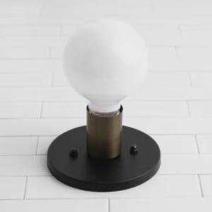 May include: A modern wall sconce featuring a large, round white light bulb. The bulb is set on a brass-colored cylinder, which is mounted on a circular black base. Two small screws secure the base. The fixture is designed for indoor use.