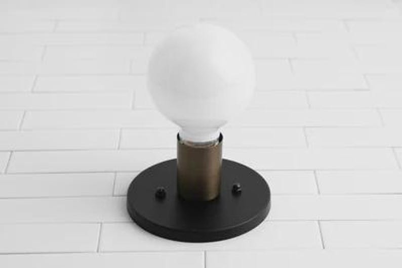 May include: A modern wall sconce featuring a large, round white light bulb. The bulb is set on a brass-colored cylinder, which is mounted on a circular black base. Two small screws secure the base. The fixture is designed for indoor use.