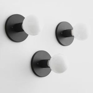 May include: Three black wall sconces with round bases and white globe-shaped light bulbs. The sconces are mounted on a white wall. The light fixtures have a minimalist design.