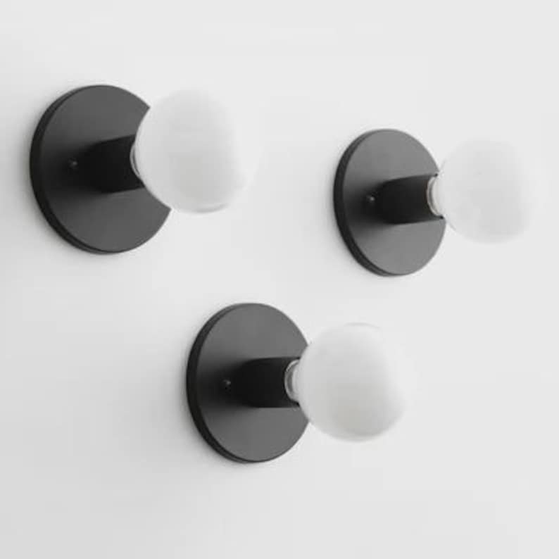 May include: Three black wall sconces with round bases and white globe-shaped light bulbs. The sconces are mounted on a white wall. The light fixtures have a minimalist design.