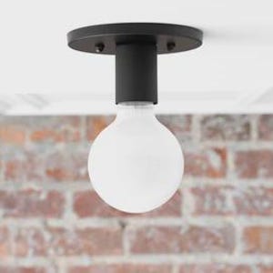 May include: A modern black ceiling light fixture with a round base and a cylindrical stem. A large, white, globe-shaped light bulb is suspended below. The background features a brick wall and a white ceiling.