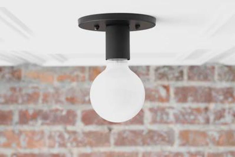 May include: A modern black ceiling light fixture with a round base and a cylindrical stem. A large, white, globe-shaped light bulb is suspended below. The background features a brick wall and a white ceiling.