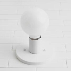 May include: A white, spherical light bulb is mounted on a white cylindrical base with a circular plate. The light fixture is set against a white, rectangular tile background. The bulb is a prominent, modern design element.