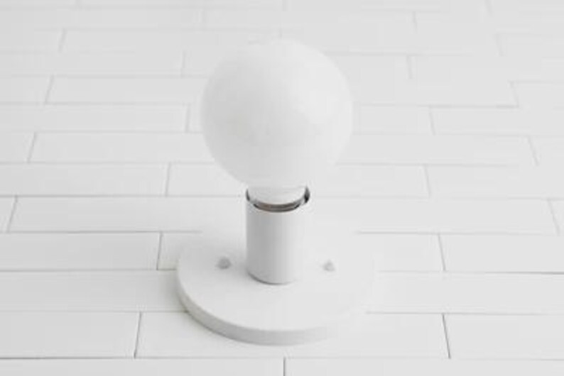 May include: A white, spherical light bulb is mounted on a white cylindrical base with a circular plate. The light fixture is set against a white, rectangular tile background. The bulb is a prominent, modern design element.