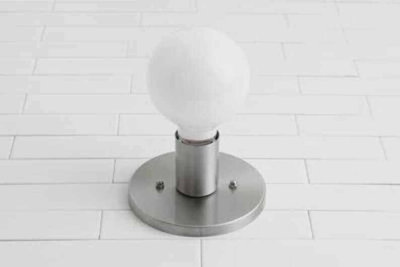 May include: A modern, round, brushed nickel wall sconce with a large, white, globe-shaped light bulb. The sconce has a circular base and a cylindrical stem. The light fixture is mounted on a white tiled wall.