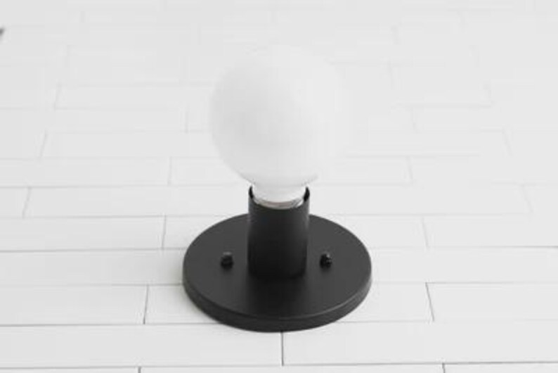 May include: A black, round wall sconce with a white, globe-shaped light bulb. The sconce has a cylindrical base and is mounted on a circular plate. The light fixture is designed for indoor use.