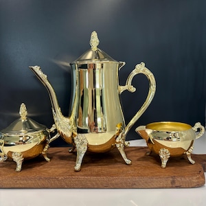 May include: A polished gold-tone tea set consisting of a tall teapot with a lid, a lidded sugar bowl, and an open creamer. Each piece features ornate, decorative feet and handles with intricate detailing, reflecting a bright, metallic finish.
