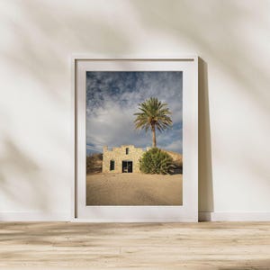 May include: Framed print of a desert landscape featuring a stone building with a dark doorway and windows. A palm tree and green shrubbery stand beside the building. The sky is cloudy with blue patches, and the ground is sandy.