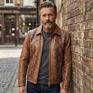 May include: A tan and brown snakeskin-patterned leather jacket with a pointed collar, long sleeves, and a front zip fastening. The jacket is worn over a dark grey t-shirt and is paired with dark blue denim jeans, featuring a silver chain necklace.