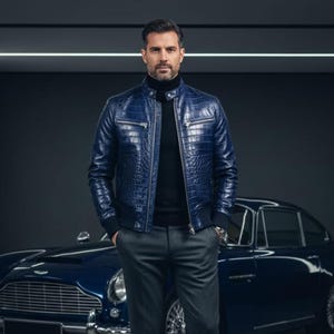 Op de afbeelding: Blau leren jack met krokodillenpatroon, ritssluiting en geribde zoom. Gedragen met een zwarte coltrui en grijze broek. Een klassieke auto is op de achtergrond te zien.