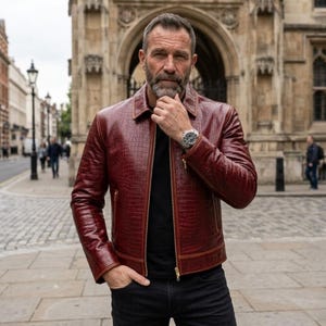 Op de afbeelding: Bordeaux leren jas met een krokodillenleer textuur. De jas heeft een ritssluiting, een kraag en een horloge om de pols. De persoon draagt een zwart shirt en een zwarte jeans. De achtergrond toont een gebouw en een straat.