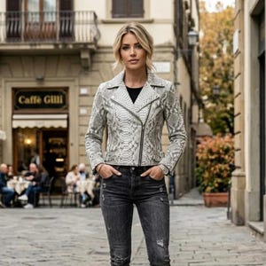 Puede incluir: Una chaqueta motera con estampado de piel de serpiente en gris claro y blanco, con cremallera negra, mangas largas y cuello de solapa, combinada con vaqueros de mezclilla gris oscuro desgastados de corte ajustado y cinco bolsillos.