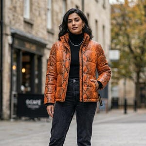 Könnte beinhalten: Eine Steppjacke mit einem leuchtend orangefarbenen und schwarzen Schlangenmuster, einem hohen Kragen und einem Frontreißverschluss. Die Jacke wird über einem schwarzen Rollkragenpullover und einer dunkelgrauen Jeans getragen, was einen Lagenlook ergibt.