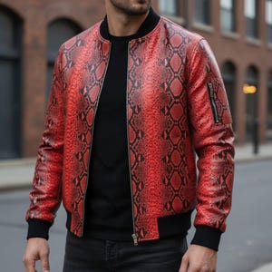 May include: A red and black snakeskin patterned bomber jacket with a black ribbed collar, cuffs, and hem. The jacket features a full-length front zipper closure and a small zippered utility pocket on the left sleeve, worn over a plain black shirt.