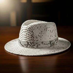 Pode incluir: Um chapéu fedora branco com um padrão de pele de cobra. O chapéu apresenta uma faixa estampada ao redor da coroa e é exibido em uma superfície de madeira. A aba do chapéu é larga e o design geral é elegante.