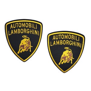 May include: Two black and gold embroidered patches featuring the Automobili Lamborghini logo. The logo includes a gold bull and text in gold. The patches are shield-shaped.