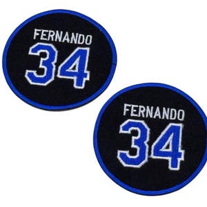 May include: Two circular patches with the name "FERNANDO" above the number "34". The text and number are in white with a blue outline, set against a black background. A blue border surrounds the black circle.