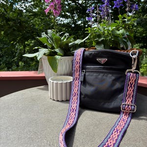 May include: A black nylon crossbody bag featuring a silver Prada logo plaque on the front, a zippered pocket, and a detachable woven shoulder strap with a repeating geometric pattern in shades of pink, purple, and light blue with silver hardware.