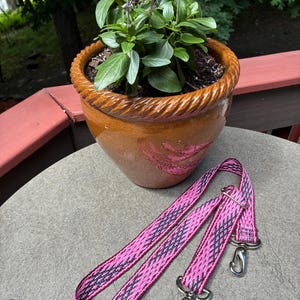 May include: A long, adjustable bag strap featuring a vibrant pink and dark gray woven geometric pattern. The strap is equipped with two polished silver-toned metal swivel lobster claw clasps at the ends, allowing for easy attachment to various bags.