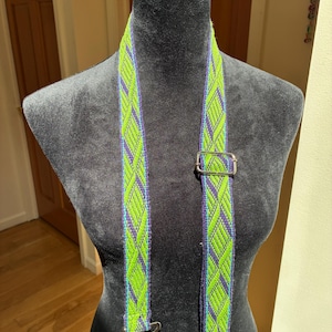 May include: A woven bag strap featuring a vibrant lime green and deep purple geometric diamond pattern. The strap includes a silver-toned metal adjustable slider buckle and two silver-toned metal swivel lobster claw clasps at the ends.