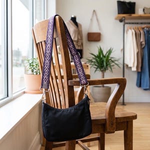 May include: A black nylon shoulder bag with a decorative strap featuring a geometric pattern in purple and white. The bag hangs on a wooden chair in a well-lit room. The strap is attached with silver-colored metal clips.