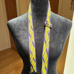 May include: A woven bag strap featuring a repeating diamond pattern in vibrant yellow and deep purple threads. The strap includes a silver-toned metal adjustable slider and two silver-toned swivel lobster claw clasps at the ends for attachment.