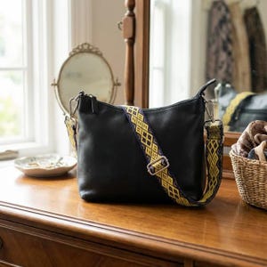 May include: Black leather crossbody bag with a patterned strap in yellow and navy blue. The bag has silver-tone hardware and a zippered top. The strap is adjustable and detachable. The bag is sitting on a wooden surface.