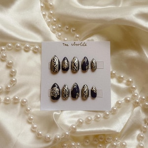 May include: A set of oval-shaped, press-on nails in a dark navy blue with gold floral and swirl designs. The nails are displayed on a white card with the text "The Isolde". A pearl necklace is draped around the nails.