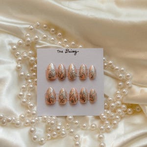 May include: A set of oval-shaped, press-on nails with a peach base and silver leaf design. The nails are displayed on a white card with the text "The Daisy". The background is a cream-colored satin fabric with a pearl necklace.